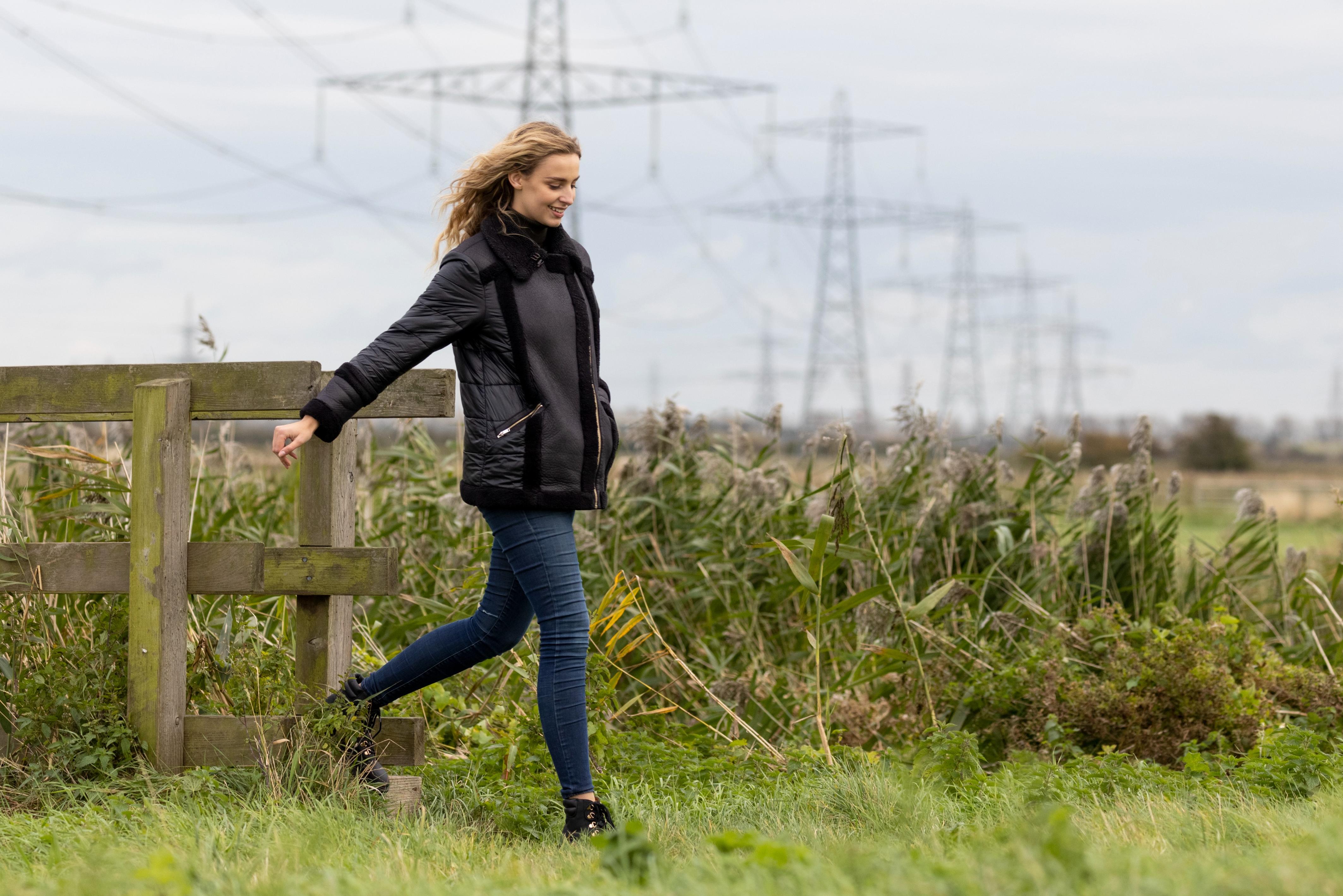 Woman walking through a grass field, looking happy, with pylons in the distance.