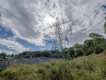 A mountainous shot looking up at an electricity pylon, with grass and trees surrounding the bottom of the pylon.