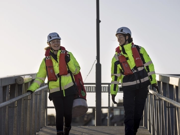 Two women walking down a bridge wearing hi vis jackets and discussing whilst looking happy.