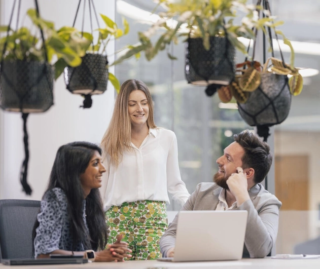 Three colleagues talking together whilst two are sat at a table and one colleague is stood up. They are all smiling.